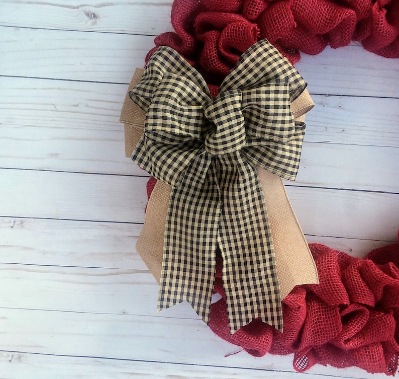 May include: A red burlap wreath with a large black and tan gingham bow. The wreath is on a white wood background.