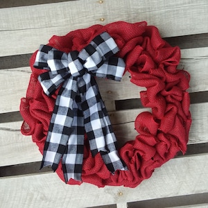 May include: A red burlap wreath with a large black and white checkered bow. The bow is tied in a knot and the tails are hanging down. The wreath is made of burlap and has a rustic look.