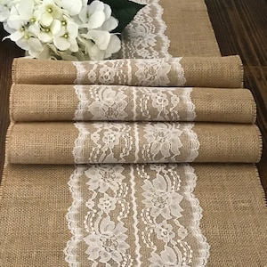 May include: A burlap table runner with a white lace trim. The runner is folded in thirds, showing the lace trim on both sides.