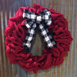 May include: A red burlap wreath with a black and white checkered bow. The wreath is made of a ruffled fabric and is perfect for a farmhouse or rustic decor style.