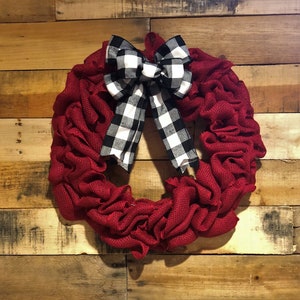May include: A red burlap wreath with a black and white checkered bow. The wreath is hanging on a wooden wall.
