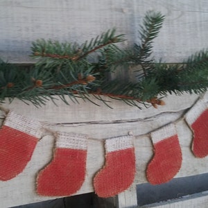May include: A garland of five red and white burlap stockings with a white string connecting them. The garland is hanging against a white wooden background with a green pine branch above it.