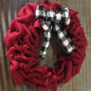 May include: A red burlap wreath with a black and white checkered bow. The wreath is hanging on a rusty metal background.
