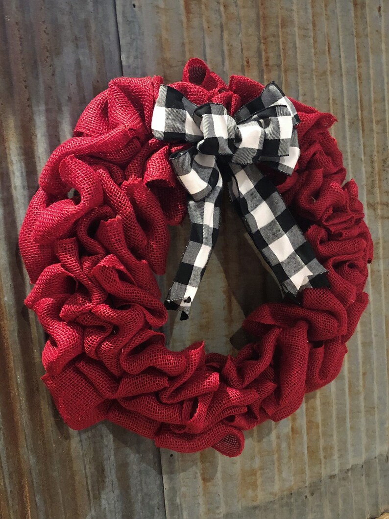 May include: A red burlap wreath with a black and white checkered bow. The wreath is hanging on a rusty metal background.