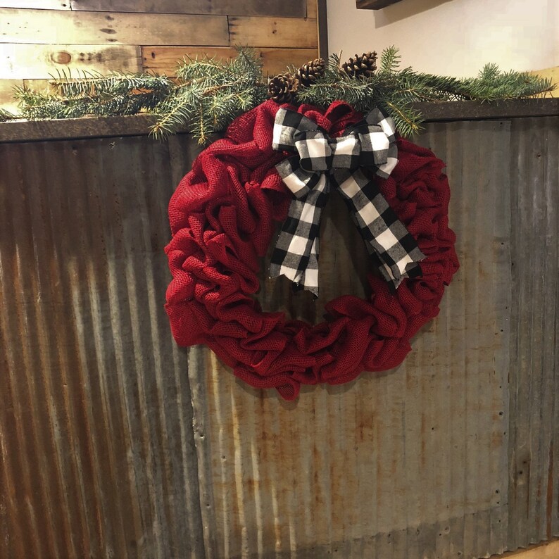 May include: A red burlap wreath with a black and white checkered bow. The wreath is hanging on a rusty metal wall.
