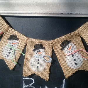 May include: A burlap banner with five snowman flags. Each flag features a white snowman with black eyes, a black hat, and a carrot nose. The snowmen are wearing colorful scarves.