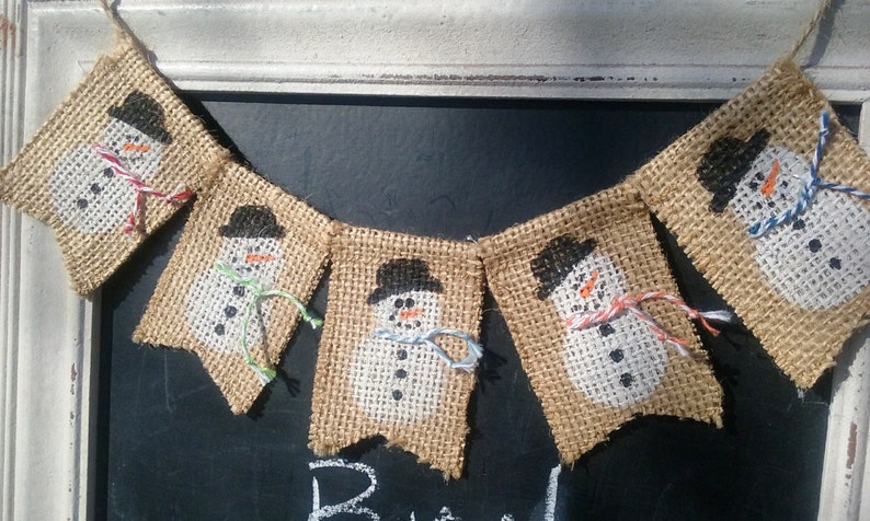 May include: A burlap banner with five snowman flags. Each flag features a white snowman with black eyes, a black hat, and a carrot nose. The snowmen are wearing colorful scarves.