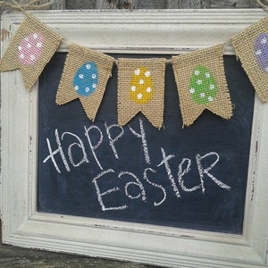 May include: A chalkboard with a white distressed frame displays the message "Happy Easter" in white chalk. A burlap banner with painted Easter eggs in pink, blue, yellow, green, and purple hangs above the chalkboard, creating a festive holiday decoration.