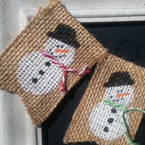 May include: A burlap banner with three painted snowmen. Each snowman has a black hat, a white face, and a black button nose. The snowmen are decorated with red, green, and white yarn.