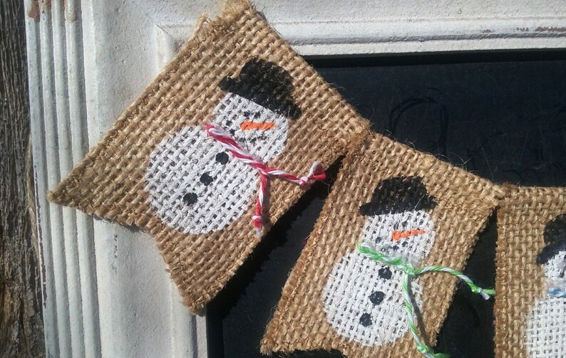 May include: A burlap banner with three painted snowmen. Each snowman has a black hat, a white face, and a black button nose. The snowmen are decorated with red, green, and white yarn.