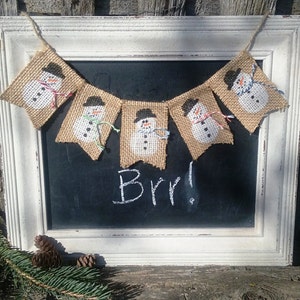 May include: A burlap banner with five snowman figures, each with a black hat and a white face. The banner is hanging on a white frame with a black chalkboard that says "Brr!"