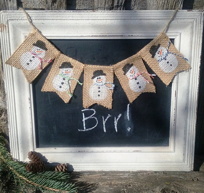 May include: A burlap banner with five snowman figures, each with a black hat and a white face. The banner is hanging on a white frame with a black chalkboard that says "Brr!"