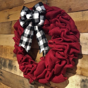 May include: A red burlap wreath with a black and white checkered bow. The wreath is made of burlap and has a ruffled texture. The bow is tied in a knot and has a long tail.