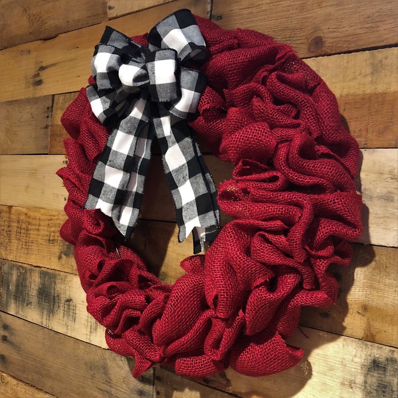 May include: A red burlap wreath with a black and white checkered bow. The wreath is made of burlap and has a ruffled texture. The bow is tied in a knot and has a long tail.