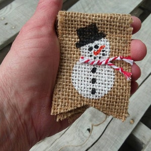 May include: A hand holding a small burlap banner with a painted snowman design. The snowman is wearing a black hat, a white scarf with red stripes, and has black eyes and a carrot nose.