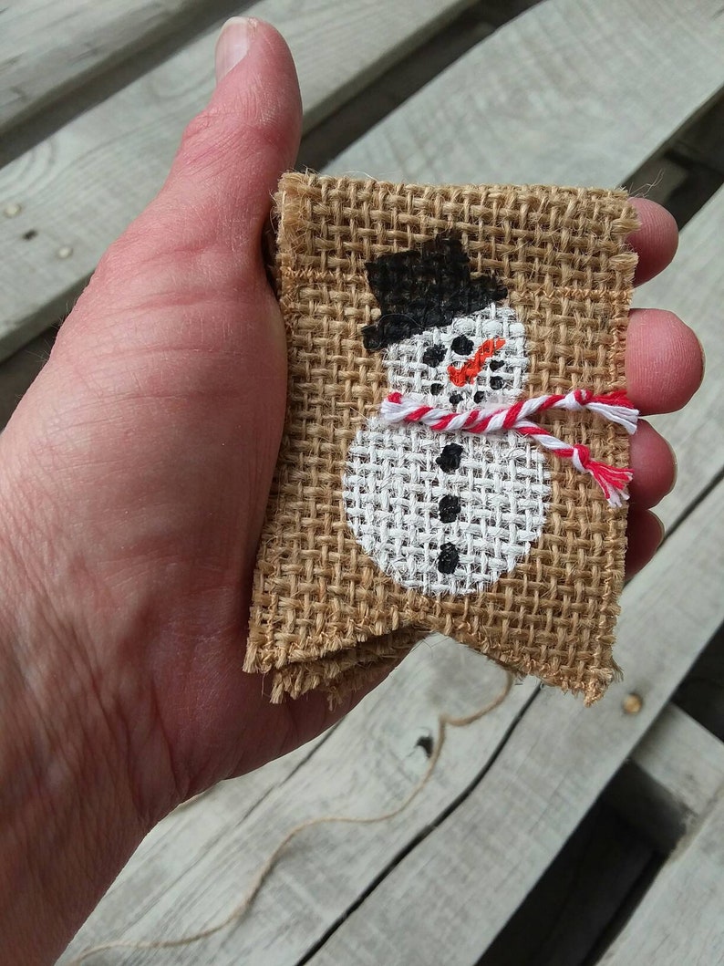 May include: A hand holding a small burlap banner with a painted snowman design. The snowman is wearing a black hat, a white scarf with red stripes, and has black eyes and a carrot nose.