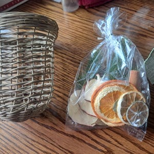 May include: A light brown woven wicker basket with an open top and a clear plastic bag containing dried orange slices, dried apple slices, green pine needles, and a cinnamon stick, tied at the top with a clear plastic twist tie.
