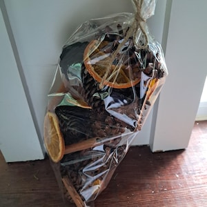 May include: A clear plastic bag tied with twine containing a collection of brown pinecones, dried orange slices, and brown cinnamon sticks. The contents are arranged together inside the transparent packaging, which is gathered and knotted at the top.