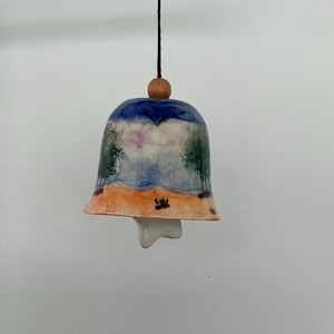 May include: Hand-painted ceramic bell with a landscape design. The bell features a blue sky, green trees, and an orange beach. A small wooden bead and black string are attached to the top. The bell has a unique, organic shape.