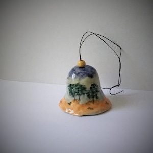 May include: Hand-painted ceramic bell with a landscape design. The bell features a blue and white sky, green trees, and an orange and tan landscape. A black string and wooden bead are attached for hanging.