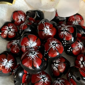 May include: A close-up of a bowl of black and red beads with white hibiscus flower designs. The beads are arranged in a random pattern and are surrounded by a white flower.