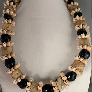 May include: A traditional Hawaiian lei necklace made with small white seashells and black beads. The shells are woven together with brown strands, creating a unique and intricate design.