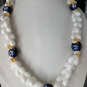 May include: A white shell lei with blue and white flower accents and gold beads. The lei is strung together with white shells and has a blue and white flower accent on each side.