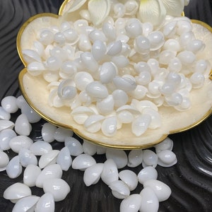 May include: A bowl filled with white seashells, with more shells scattered on a black surface. The shells are smooth and have a pearlescent sheen.
