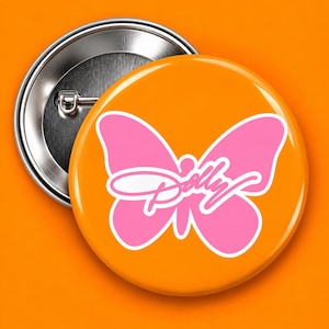 May include: A round pin-back button featuring a bright orange background with a large, solid pink butterfly graphic centered on the front. The word Dolly is written in a white cursive script font across the center of the pink butterfly shape.