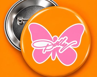May include: A round pin-back button featuring a bright orange background with a large, solid pink butterfly graphic centered on the front. The word Dolly is written in a white cursive script font across the center of the pink butterfly shape.