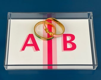 Könnte beinhalten: Zwei aufklappbare Metallarmreifen, einer mit grüner und einer mit rosa Emaille-Einlage, liegen auf einem rechteckigen, durchsichtigen Acryltablett. Das Tablett enthält eine weiße Einlage mit einem rosa vertikalen Streifen sowie den Buchstaben A und B.