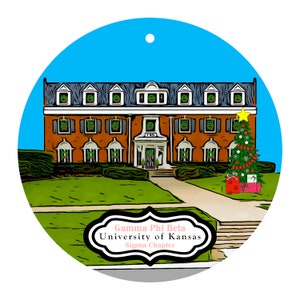 May include: A round Christmas ornament featuring a brick building with green wreaths on the windows and a decorated Christmas tree. The ornament has a blue sky background and text that reads "Gamma Phi Beta, University of Kansas, Sigma Chapter."
