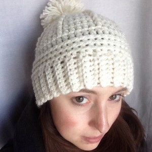May include: A white crocheted beanie with a white pom pom on top. The beanie is worn by a person with brown hair.