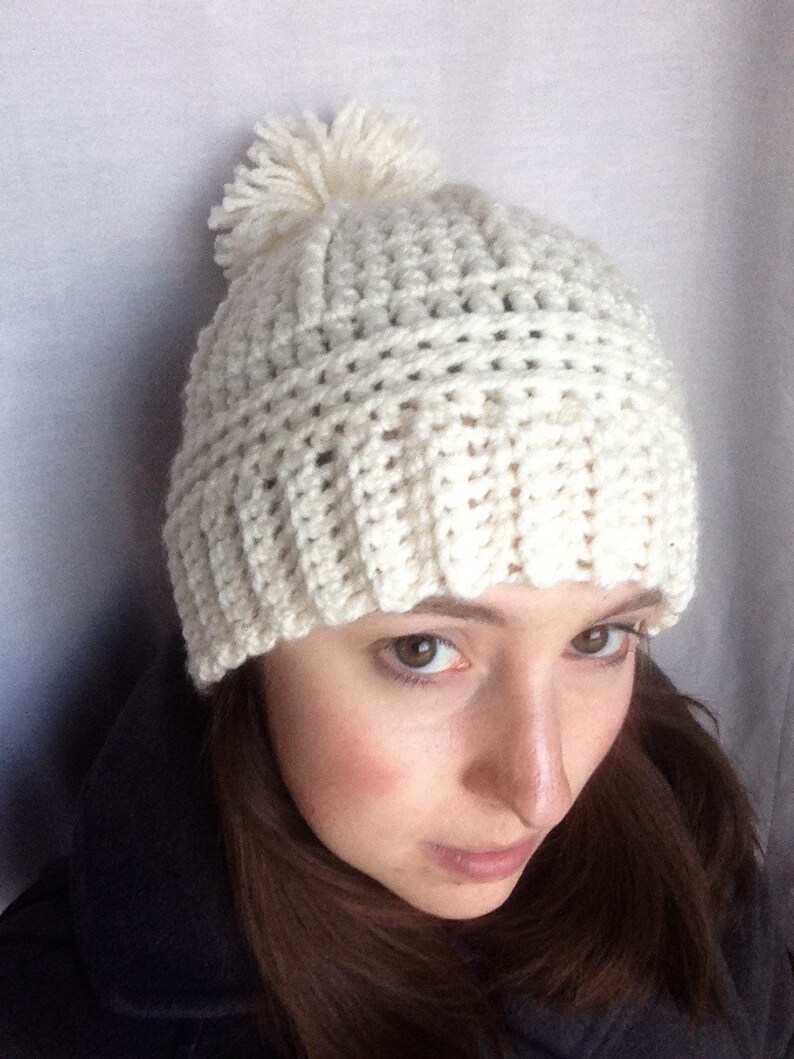 May include: A white crocheted beanie with a white pom pom on top. The beanie is worn by a person with brown hair.