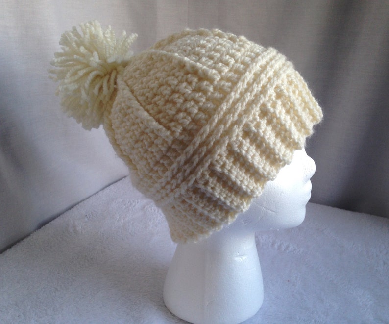 May include: A cream-colored crocheted beanie with a white pom-pom on top. The beanie is made with a textured stitch pattern.