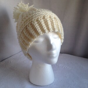 May include: A crocheted beanie hat in a cream color with a fluffy pom pom on top.