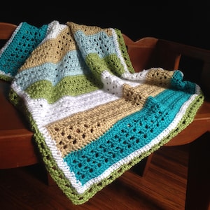 May include: A crocheted baby blanket in shades of teal, green, white, and tan. The blanket is made with a variety of stitches and has a fringed edge.