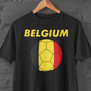 Puede incluir: Camiseta negra con la palabra "BELGIUM" en amarillo sobre un diseño de balón de fútbol. El balón incorpora los colores negro, amarillo y rojo de la bandera belga. La camiseta está colgada en una percha de madera contra una pared de ladrillos gris oscuro.