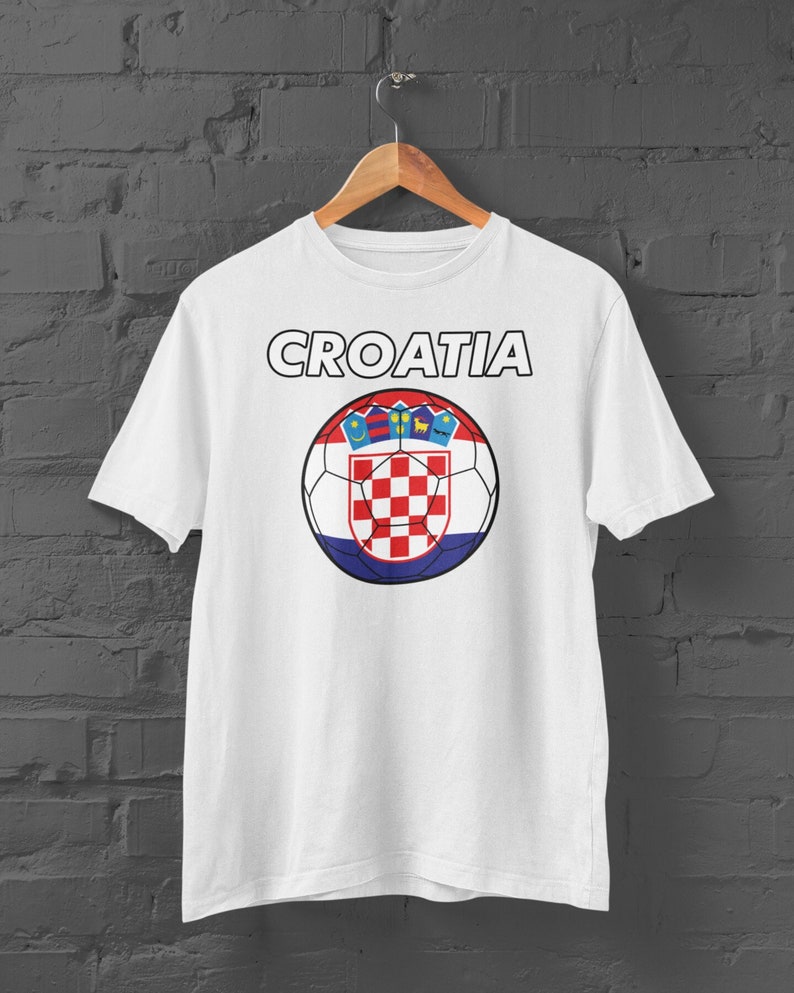 May include: White t-shirt with the word "CROATIA" above a soccer ball design. The soccer ball features the Croatian coat of arms and flag colors: red, white, and blue. The shirt is hanging on a wooden hanger against a dark brick wall.