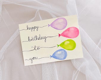 May include: A handwritten card with the words "happy birthday to you" and four watercolor balloons in purple, pink, green, and blue.