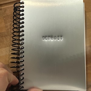 以下が含まれることがあります： 黒いスパイラル綴じの銀色のノート。表紙の中央には「DETROIT」の文字が刻印されています。ノートは木製の表面に置かれています。