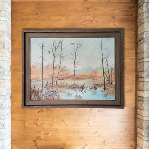 May include: A framed landscape painting depicting a serene marsh scene. Bare trees, a small boat, and flying ducks are visible against a backdrop of muted colors. The artwork is mounted on a wooden wall.