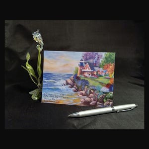 May include: A rectangular postcard titled Guided by Peace by Diane L. Wolf featuring a lighthouse on a rocky coast, a green glass sculpture of a flower, and a silver ballpoint pen. The scene depicts a coastal landscape with blue waves.