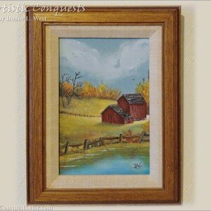 May include: A framed painting of a rural landscape with a red barn and a pond. The painting is in a wooden frame with a light brown finish. The painting is titled "Artistic Conquests" by Duane L. West.