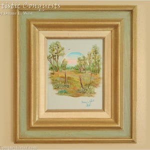 May include: Framed landscape painting with a rural scene. The artwork features a field, trees, and a small building under a blue and pink sky. The frame is gold and light blue, with the text "Artistic Conquests" at the top.