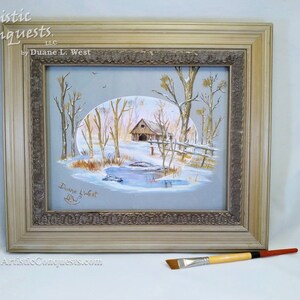 May include: A framed painting of a winter scene with a cabin, trees, and a pond. The painting is signed "Duane L. West" in the bottom left corner. The frame is a light brown color with a decorative border. The painting is titled "Artistic Conquests, LLC by Duane L. West".