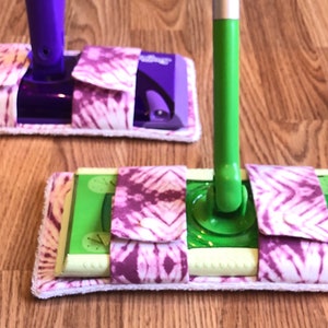 May include: Two mops with green and purple handles and tie-dye patterned covers. The covers are white with pink and purple tie-dye patterns.