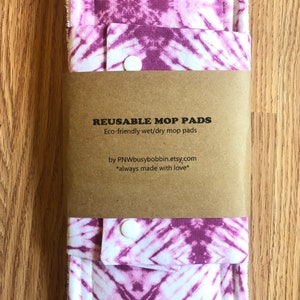 May include: Two purple and white tie-dye reusable mop pads with snap closures. The pads are packaged in a brown paper envelope with the text "REUSABLE MOP PADS" and "Eco-friendly wet/dry mop pads" printed on it. The text "by PNWbusybobbin.etsy.com" and "*always made with love*" is also printed on the envelope.