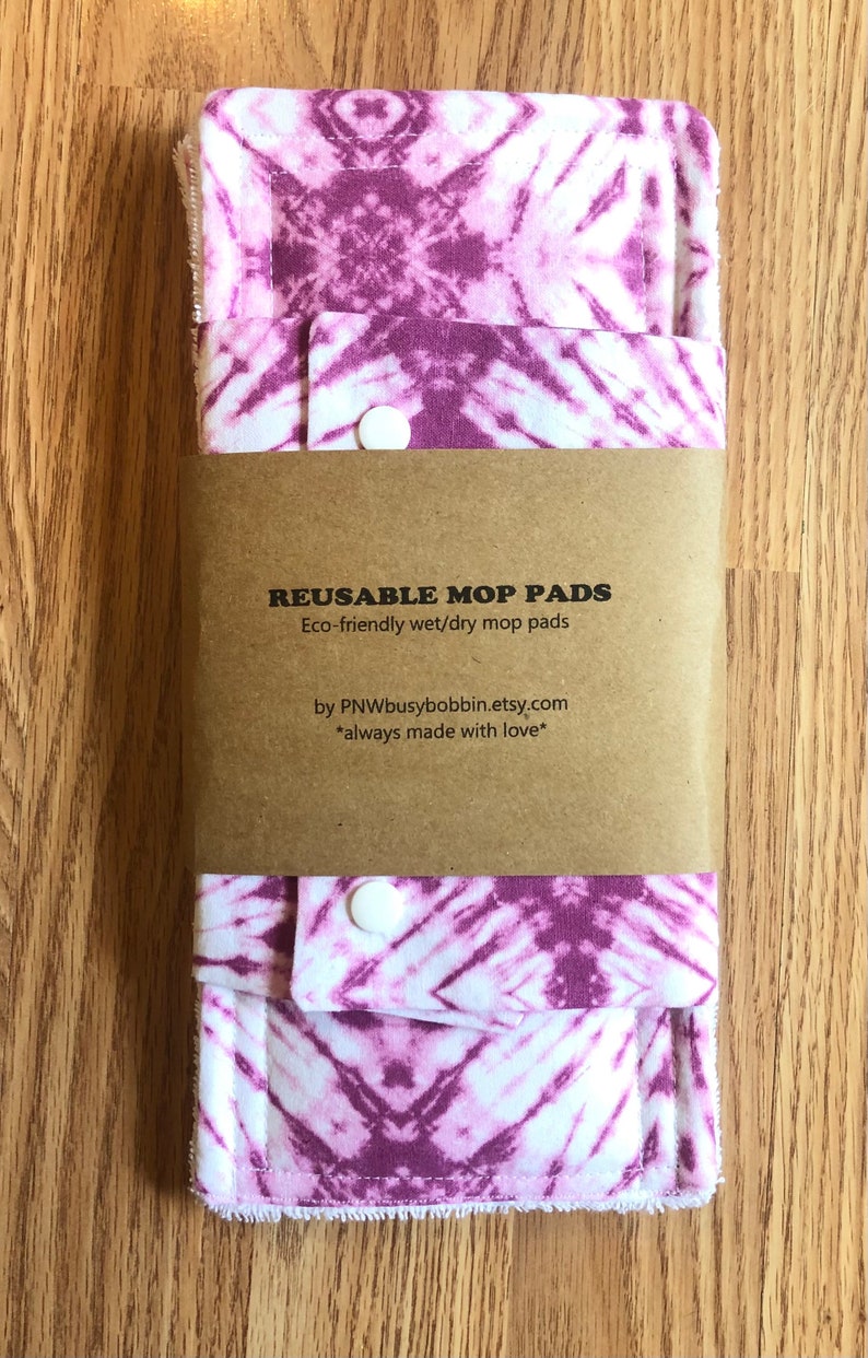 May include: Two purple and white tie-dye reusable mop pads with snap closures. The pads are packaged in a brown paper envelope with the text "REUSABLE MOP PADS" and "Eco-friendly wet/dry mop pads" printed on it. The text "by PNWbusybobbin.etsy.com" and "*always made with love*" is also printed on the envelope.