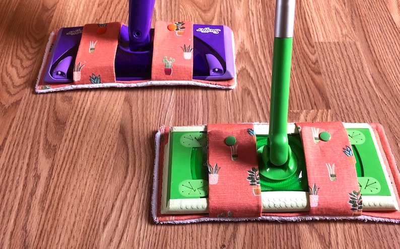 May include: Two Swiffer mop heads with custom fabric covers. The covers are orange with a green and white floral pattern. One mop head is purple and the other is green.